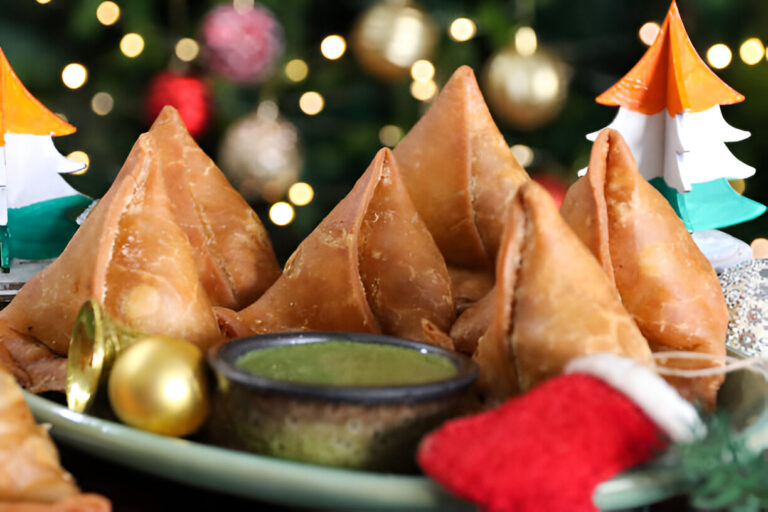 Plate of golden-brown samosas served with green chutney and decorative holiday trees in the background.
