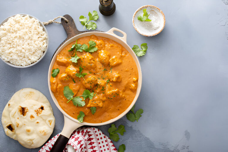 A serving of chicken curry garnished with fresh cilantro, accompanied by a bowl of white rice, a piece of naan bread, and a small dish of salt, all on a light blue background.