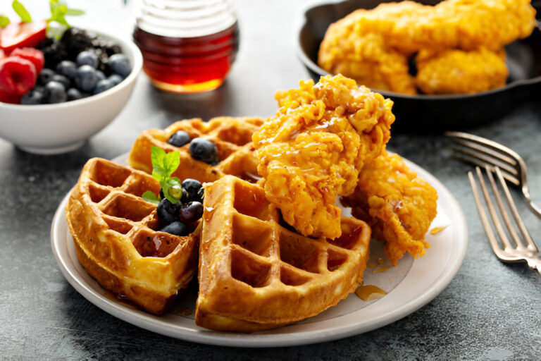 A plate with crispy fried chicken and fluffy waffles drizzled with syrup, garnished with fresh blueberries and mint leaves, alongside a bowl of mixed berries.