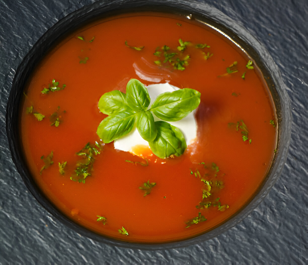 A bowl of vibrant tomato soup topped with fresh basil and cream, placed on a textured black surface.