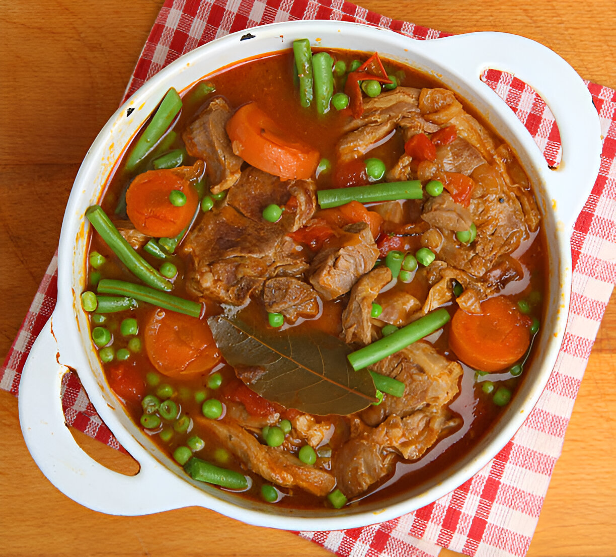 A delicious beef stew with green beans, peas, carrots, and bay leaves in a white casserole dish on a red checkered cloth.