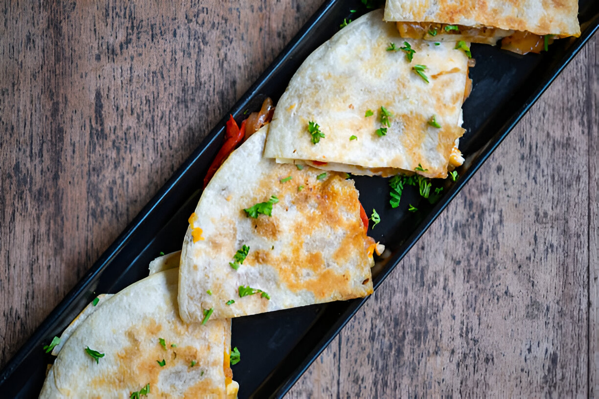 A plate of freshly made quesadillas filled with vegetables, garnished with parsley, arranged on a dark serving platter against a wooden background.