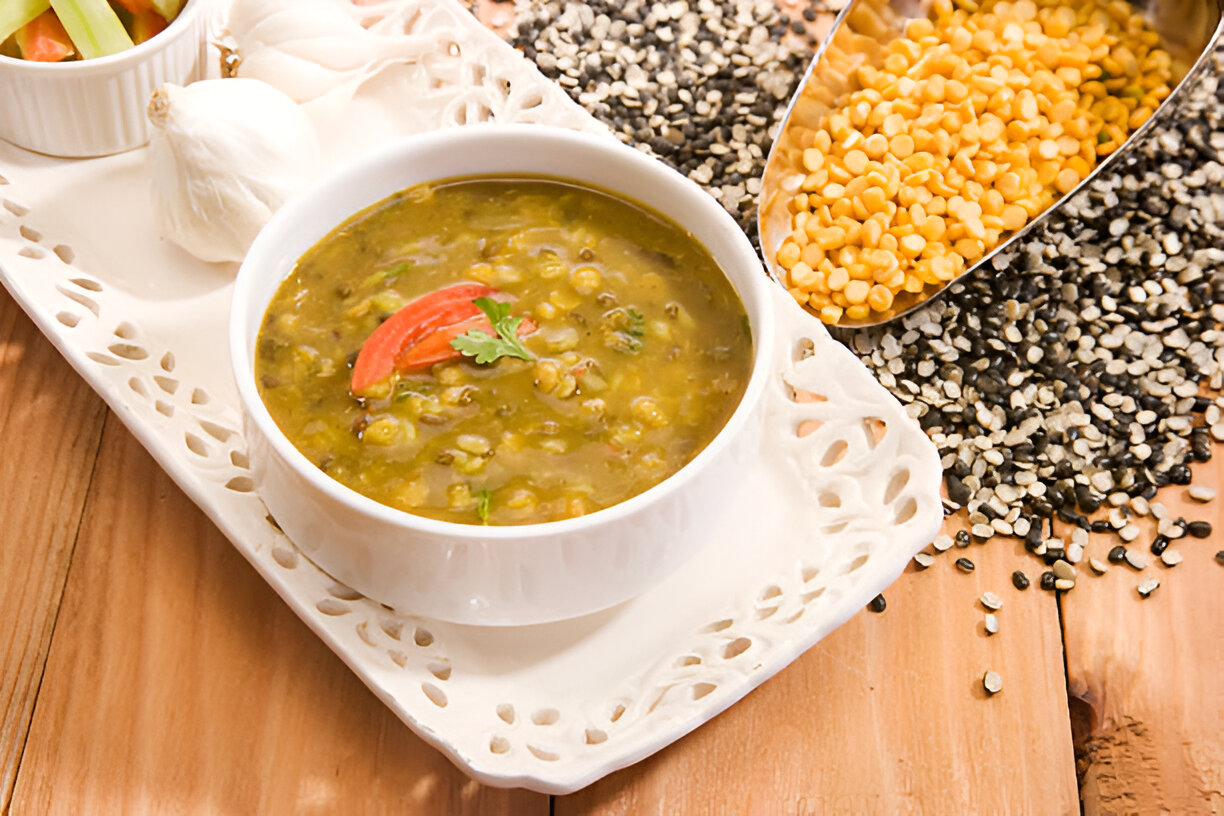 A bowl of green lentil soup garnished with tomato slices and coriander, served on a decorative tray with garlic and yellow lentils in the background.