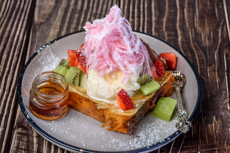 A delectable dessert consisting of thick toast topped with ice cream, fresh fruits, and pink cotton candy, served with syrup on a wooden table.
