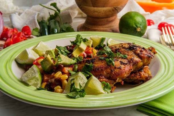A plate of grilled chicken served with lime wedges and a colorful bean salad consisting of corn, tomatoes, and avocado.