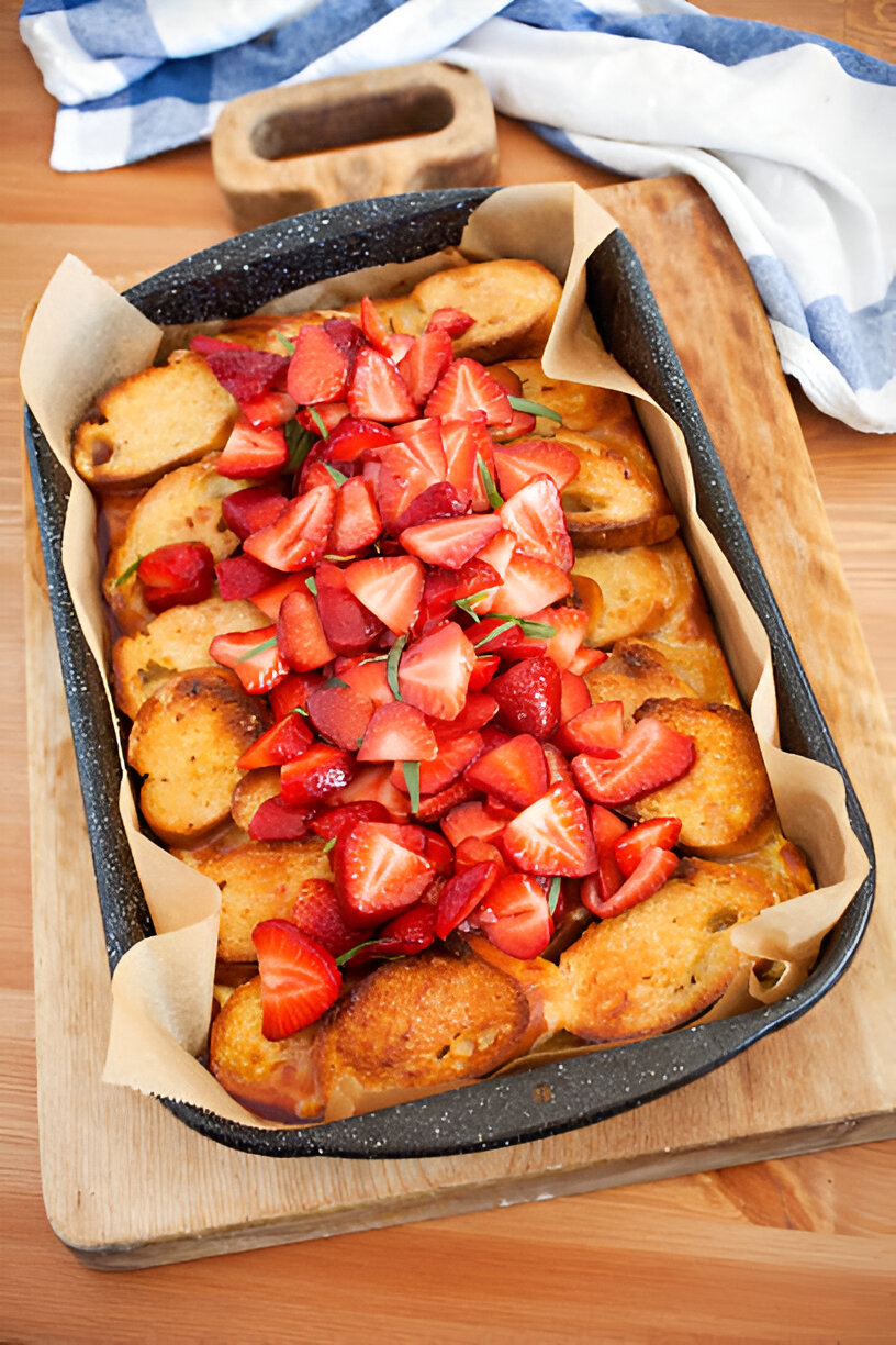 Baked dessert topped with fresh strawberries and green herbs in a rectangular tray on a wooden surface.