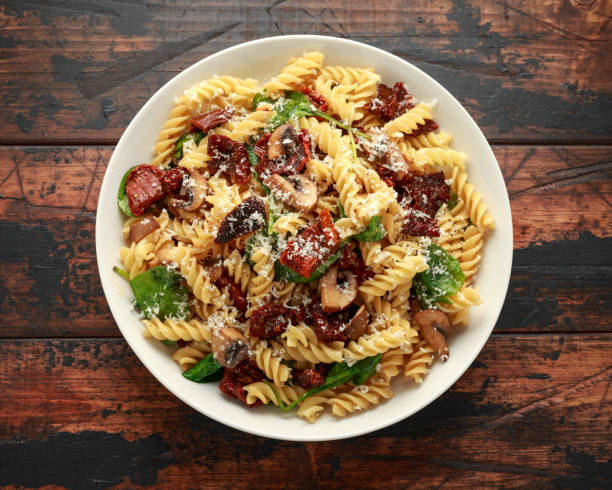 A bowl of pasta with sun-dried tomatoes, mushrooms, and spinach on a wooden table.