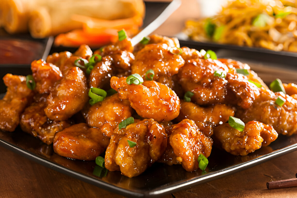 A plate of crispy chicken pieces glazed with a shiny sauce, garnished with chopped green onions, accompanied by spring rolls and noodles in the background.