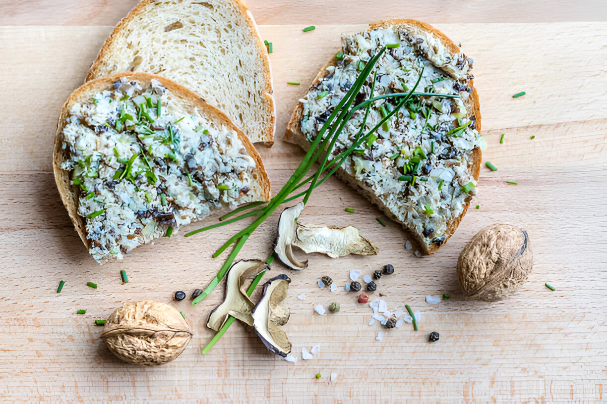 Two slices of bread spread with a creamy topping, garnished with chopped chives, surrounded by mushrooms, spices, and nuts on a wooden surface.