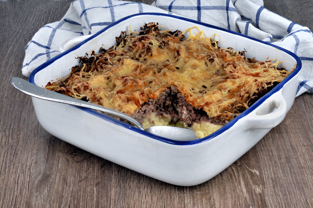 A casserole dish filled with a layered meat and cheese dish, with a silver spoon resting beside it.