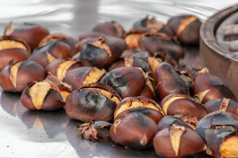 Roasted chestnuts piled on a metallic surface, some shells cracked open revealing the yellow nut inside.