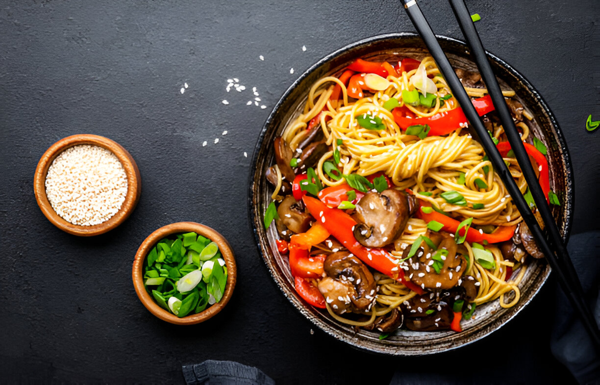 A bowl of stir-fried noodles with colorful bell peppers, mushrooms, and green onions, surrounded by small bowls of sesame seeds and chopped green onions.