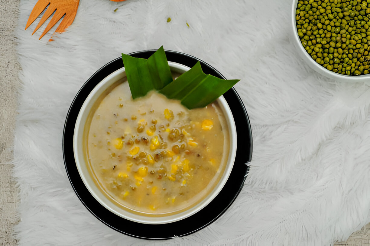 A bowl of creamy corn and mung bean soup garnished with pandan leaves, surrounded by ingredients on a soft white surface.