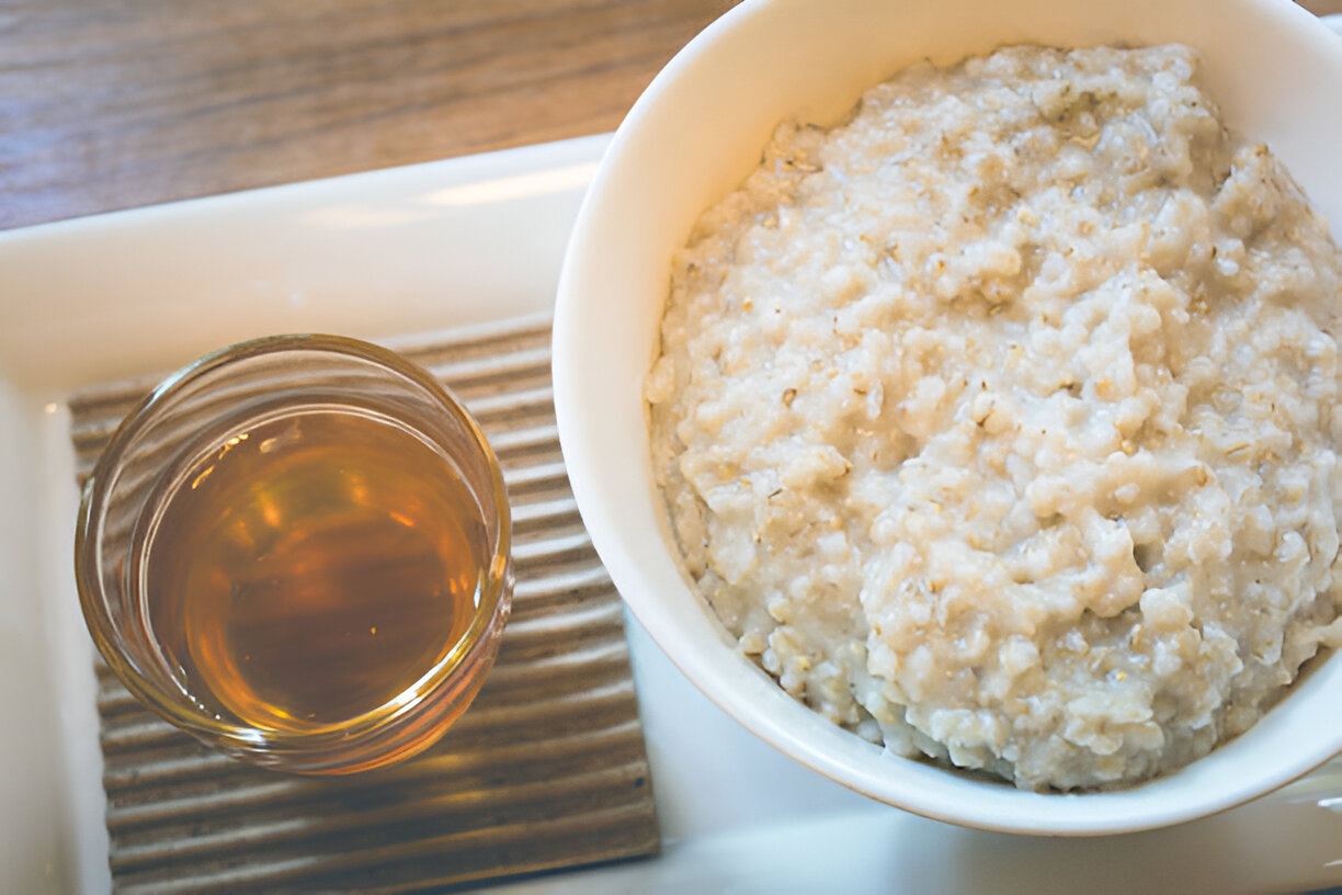 A bowl of creamy oatmeal served with a glass of syrup on a wooden table.