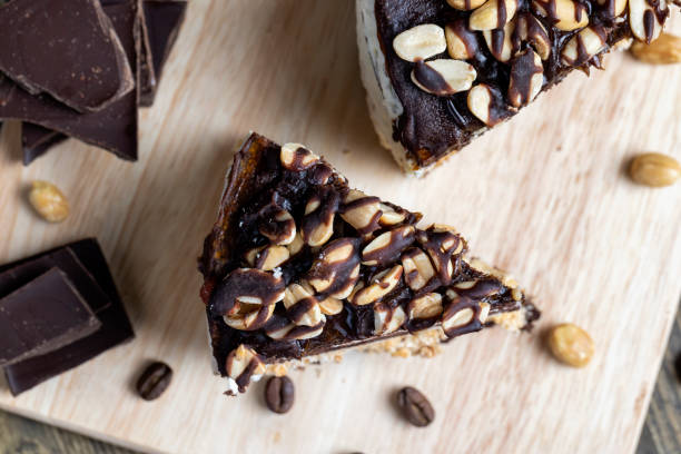 A slice of chocolate cake topped with nuts on a wooden board, surrounded by chocolate pieces and coffee beans.