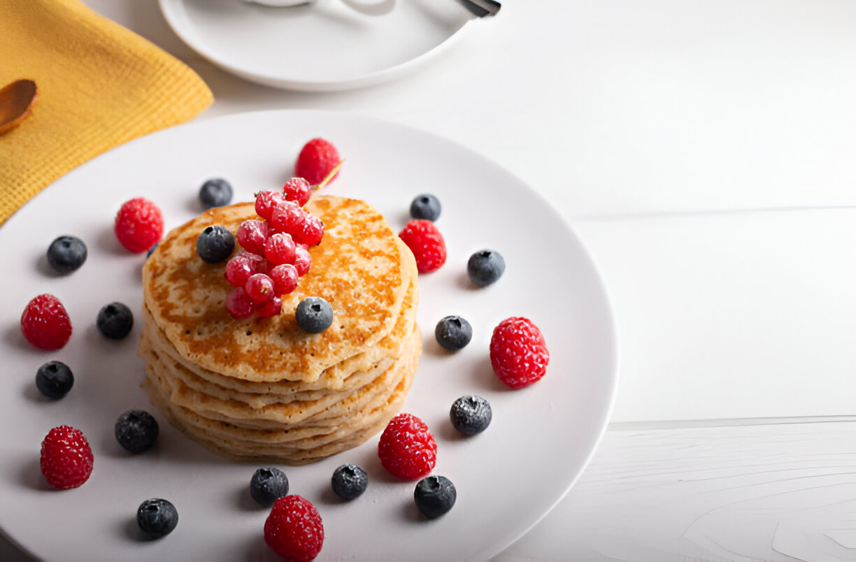A stack of pancakes topped with fresh red currants and surrounded by raspberries and blueberries on a white plate.