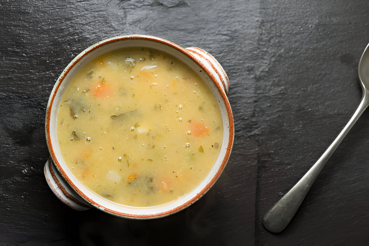 A bowl of soup with vegetables on a dark slate surface, accompanied by a silver spoon.