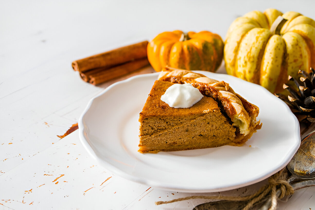 A slice of pumpkin pie topped with whipped cream on a white plate, surrounded by pumpkins and cinnamon sticks.