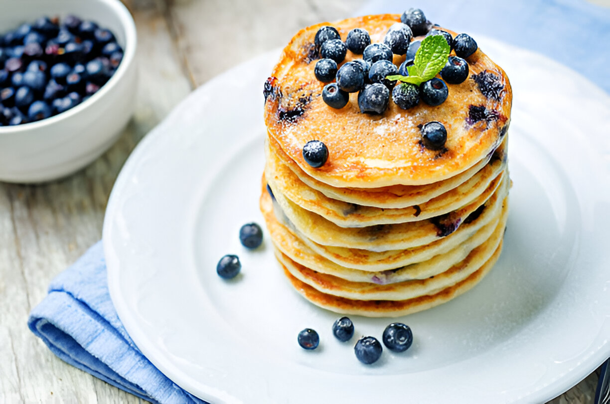 A stack of fluffy pancakes topped with fresh blueberries and a sprig of mint on a white plate, accompanied by a bowl of blueberries.
