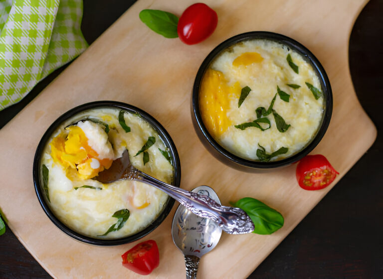 Two bowls of creamy baked egg dish with basil on a wooden board, accompanied by cherry tomatoes and a spoon