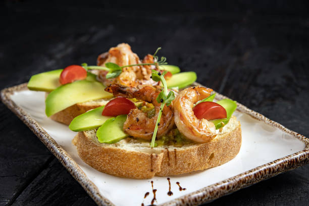 Open-faced sandwich topped with grilled shrimp, avocado slices, cherry tomatoes, and microgreens on a white plate.