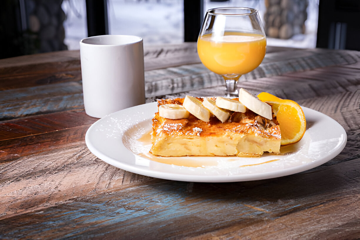 A delicious slice of dessert topped with banana slices and a wedge of orange, served on a white plate with a glass of orange juice and a mug in the background.