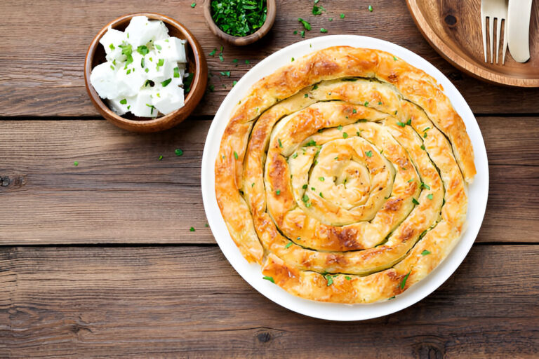 A spiral pastry dish garnished with green herbs on a white plate, accompanied by bowls of cheese and chopped herbs.