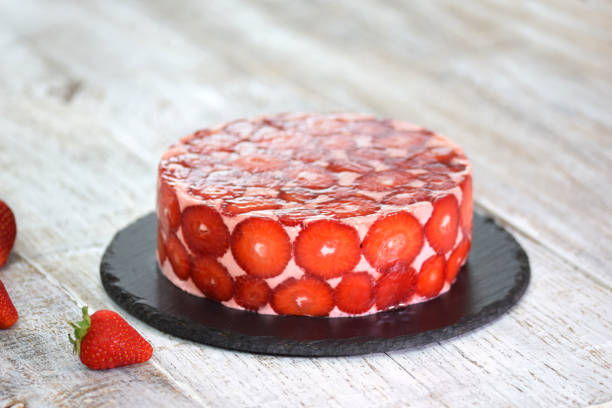 A round strawberry cake set on a black plate, surrounded by fresh strawberries on a wooden surface.