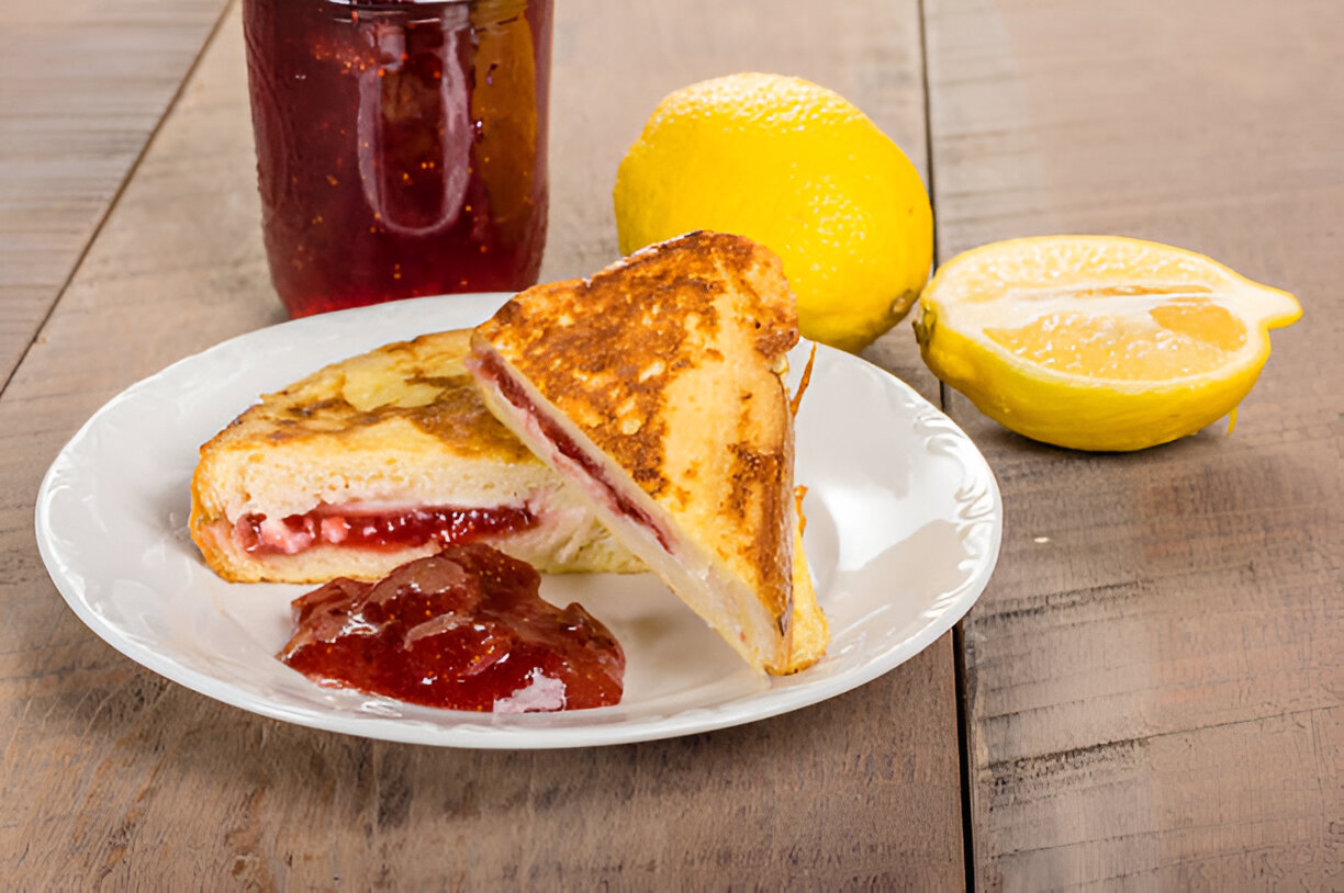 A plate with a toasted sandwich filled with jam, accompanied by a jar of jam and fresh lemons on a rustic wooden table.