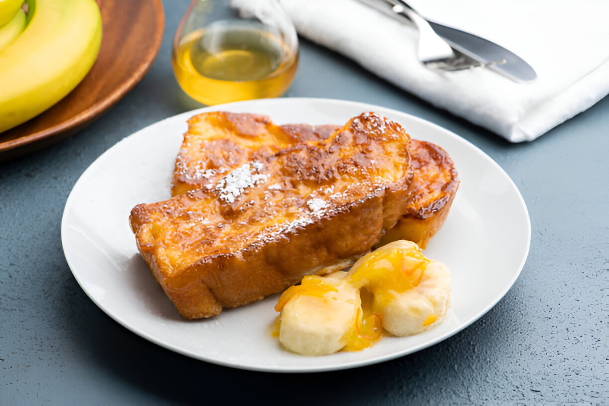 A plate of French toast topped with powdered sugar and served with banana slices and orange sauce.