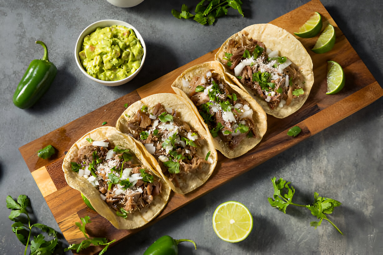 Three delicious tacos topped with cilantro and onions, served on a wooden board alongside guacamole and lime wedges.