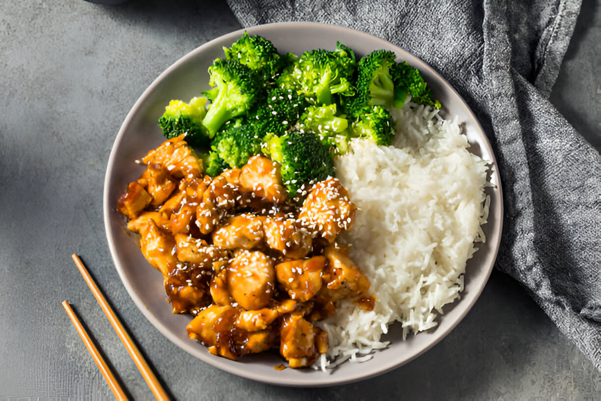A bowl of sesame chicken, steamed broccoli, and white rice served with chopsticks.