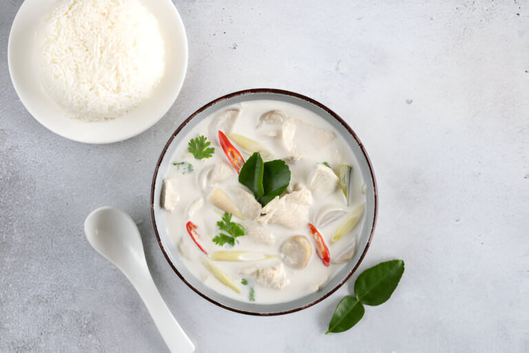 A bowl of creamy soup with chicken, herbs, and vegetables, accompanied by a side of steamed rice.