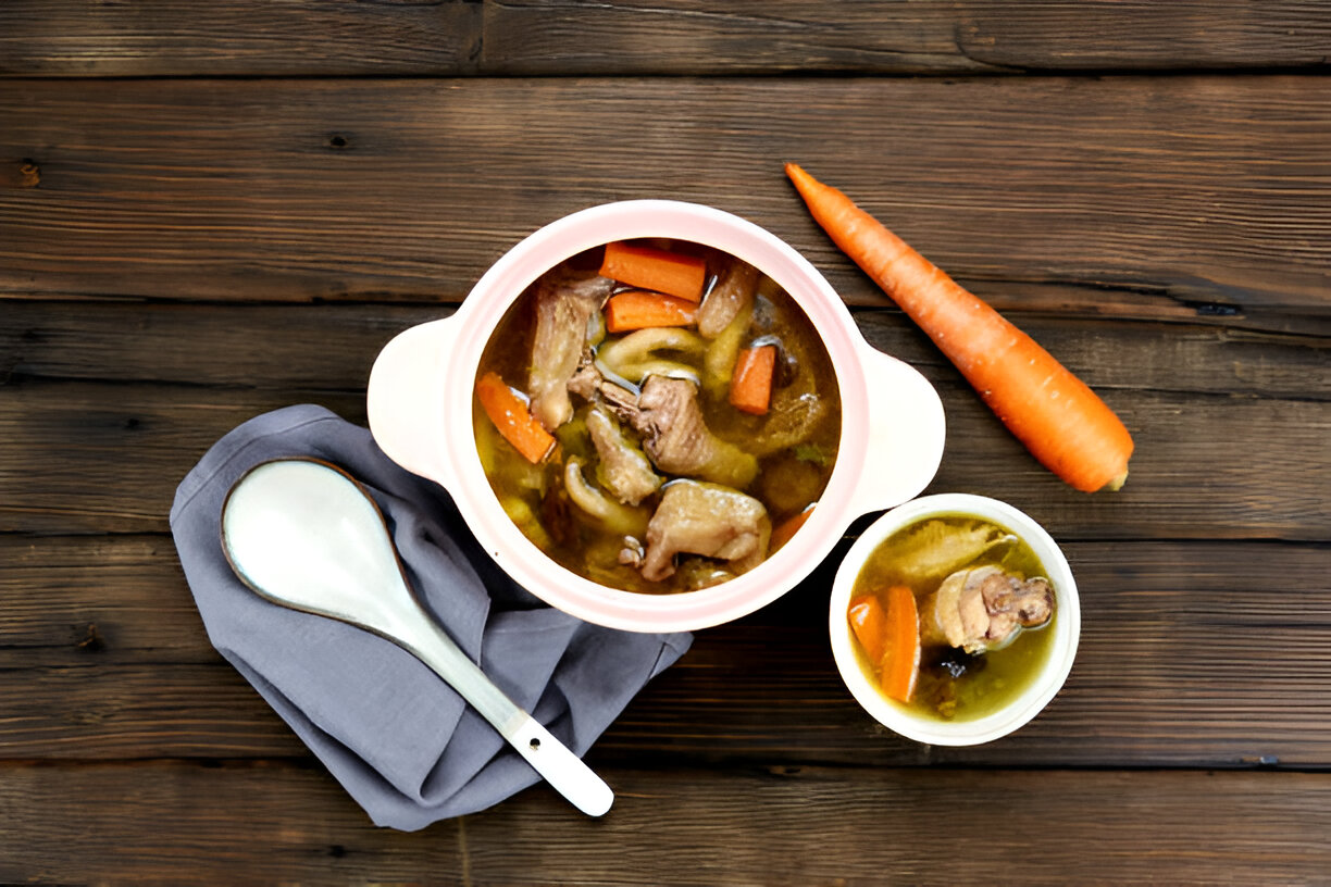 A bowl of hearty chicken soup with carrots, served in a white pot, accompanied by a small bowl and a spoon on a gray cloth, against a wooden background.