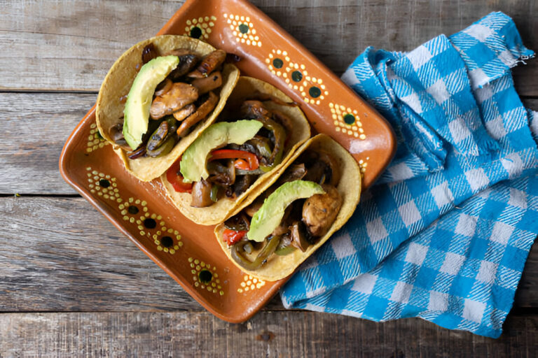 Three chicken fajita tacos served on a decorative plate with sliced avocado, placed on a wooden table