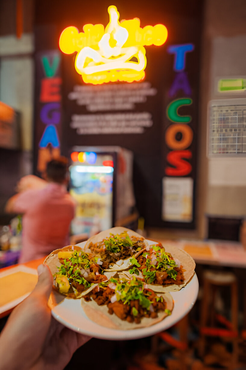A close-up of a plate of tacos topped with fresh vegetables held in a hand, with a colorful restaurant background.