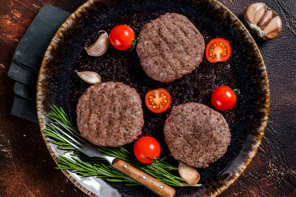 A close-up of three raw burger patties on a dark plate, garnished with cherry tomatoes, garlic cloves, and fresh rosemary.
