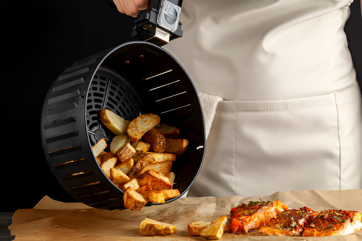 A person in an apron pouring crispy potato wedges from an air fryer basket onto a wooden surface next to a piece of cooked chicken.
