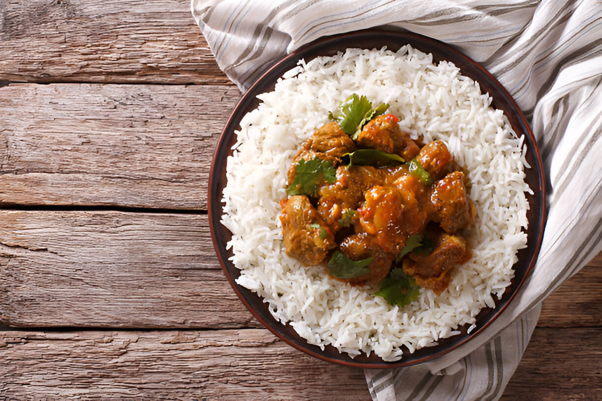 A bowl of rice topped with flavorful meat curry and garnished with fresh herbs, placed on a wooden surface.