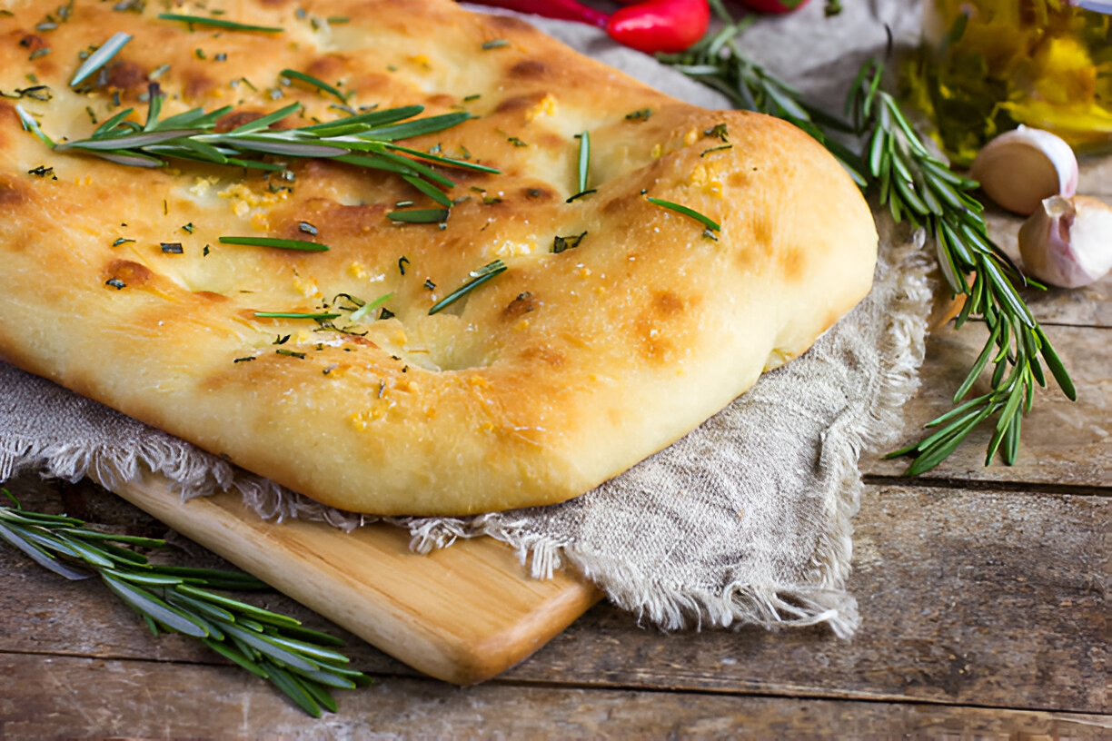 Freshly baked focaccia bread topped with herbs, placed on a wooden cutting board.