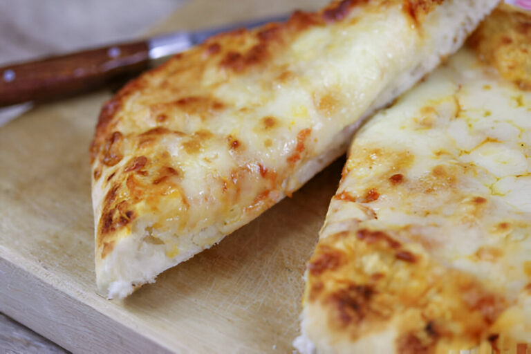 A freshly baked cheese flatbread cut into slices on a wooden board with a knife in the background.