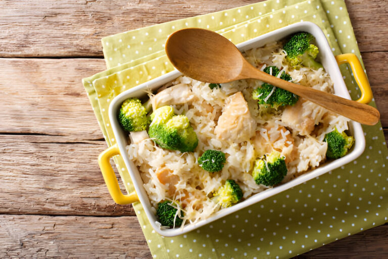 A creamy chicken and broccoli casserole in a white dish with a wooden spoon, placed on a green polka dot cloth on a rustic wooden table.
