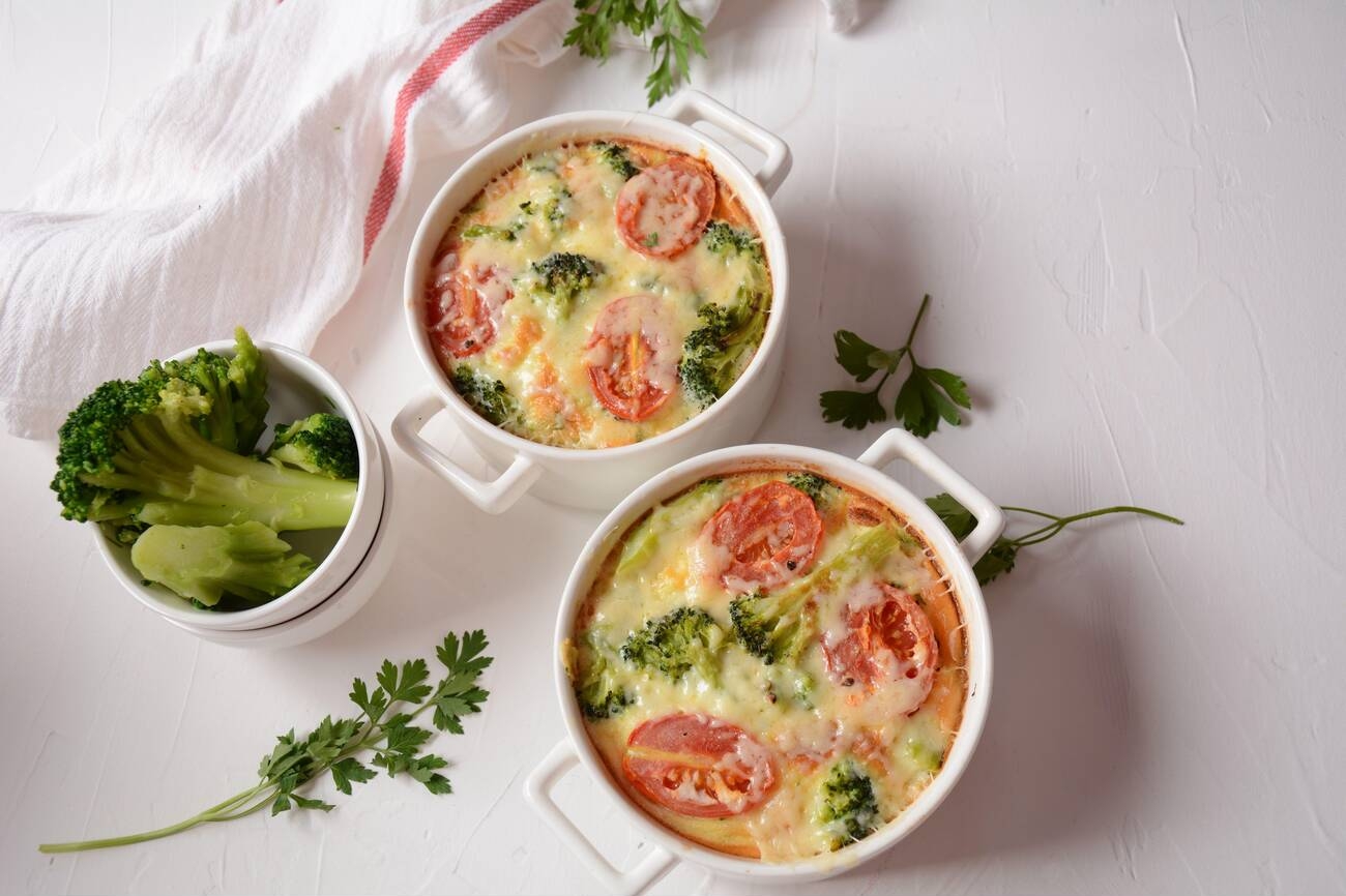 Two white ramekins filled with baked broccoli and tomato dish, accompanied by a small bowl of broccoli and fresh parsley on a light surface.