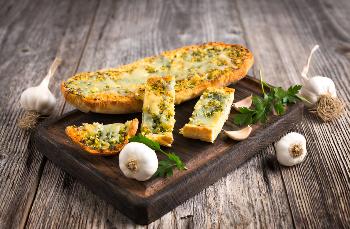 A freshly baked garlic bread topped with herbs, served on a wooden board with garlic cloves and parsley.