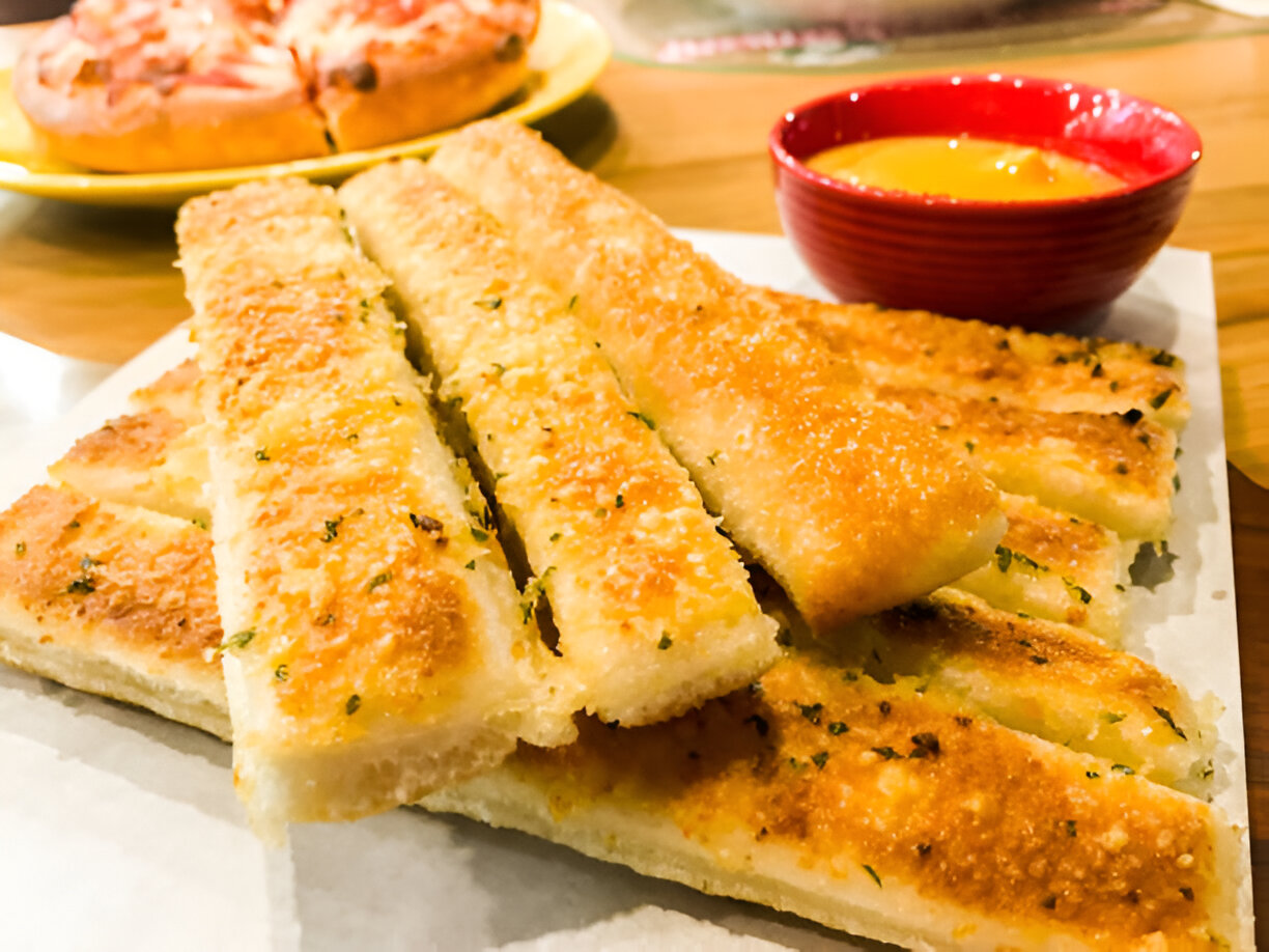 A plate of golden garlic bread sticks served with a red bowl of dipping sauce and a pizza in the background.