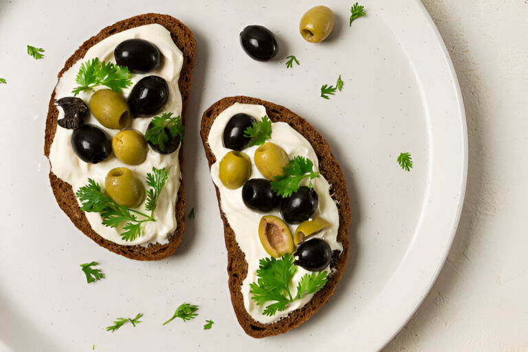 Two slices of dark bread topped with cream cheese, black and green olives, and fresh cilantro on a white plate