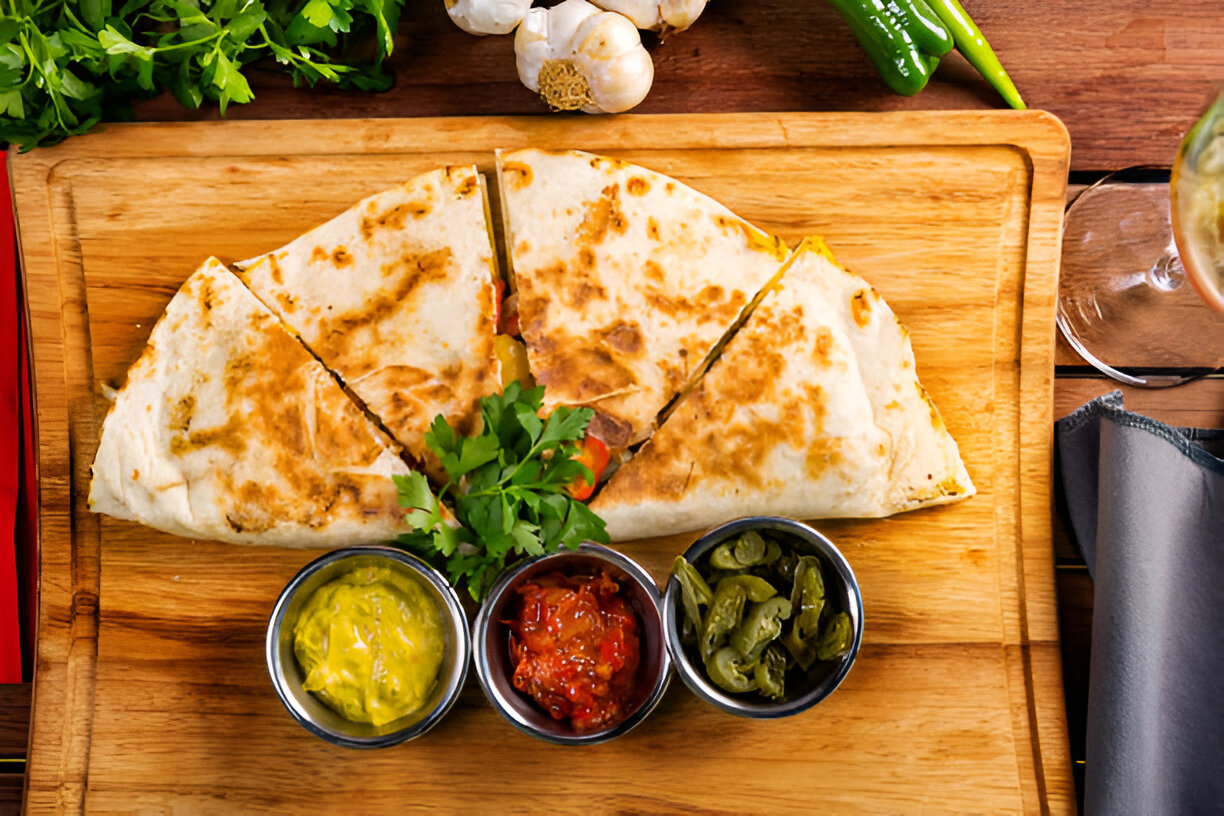 A delicious quesadilla cut into wedges on a wooden board, accompanied by dips of guacamole, salsa, and jalapeños.