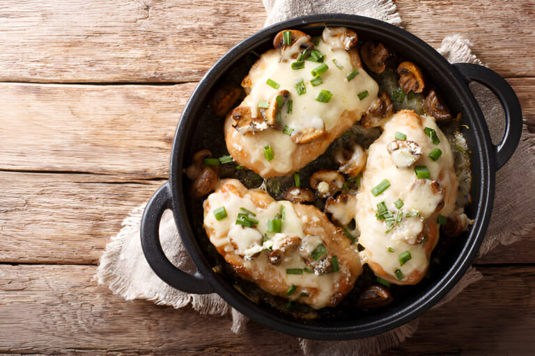 A cast iron dish containing baked chicken topped with melted cheese and sautéed mushrooms, garnished with chopped green onions, on a rustic wooden surface.