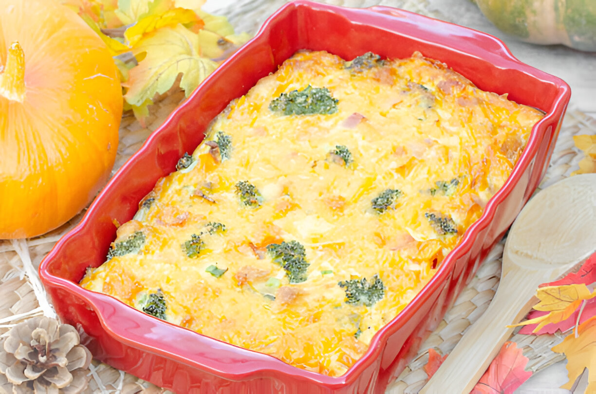 A red casserole dish filled with baked broccoli and cheese, surrounded by autumn-themed decorations including a small pumpkin and colorful leaves.