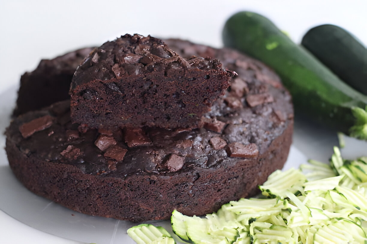 A chocolate zucchini cake with pieces of chocolate on top, alongside sliced zucchini and whole zucchini in the background.